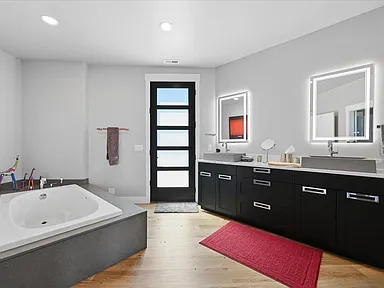 A freestanding soaking tub sits beside a dark wood double vanity with backlit mirrors