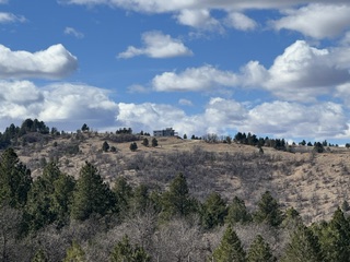 A distant hillside view shows 2111 Vantage Circle atop a tree-lined ridge under a partly cloudy sky.