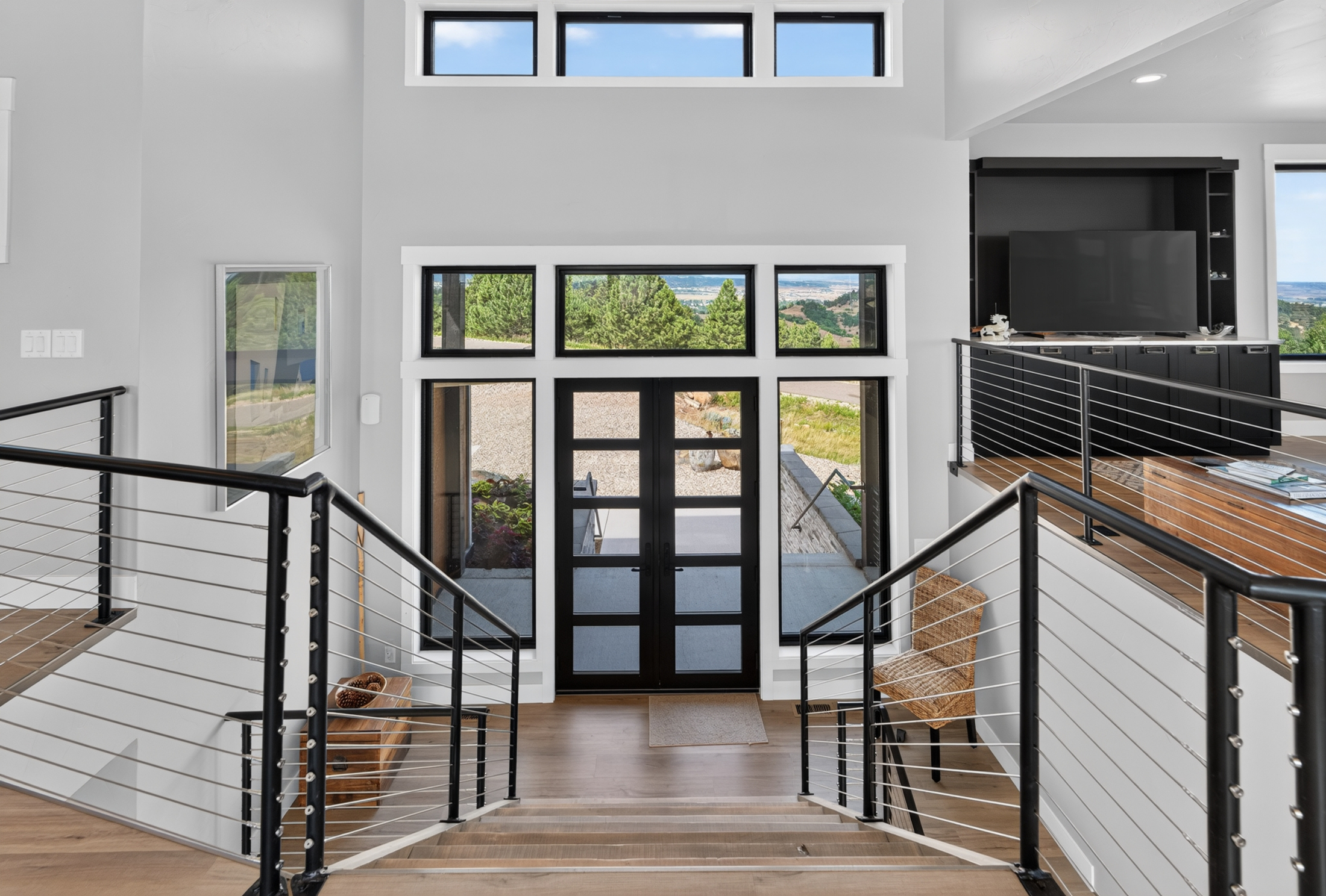 Black-framed double entry doors are flanked by tall windows, with a woven chair and pinecone-filled bowl in the foyer.